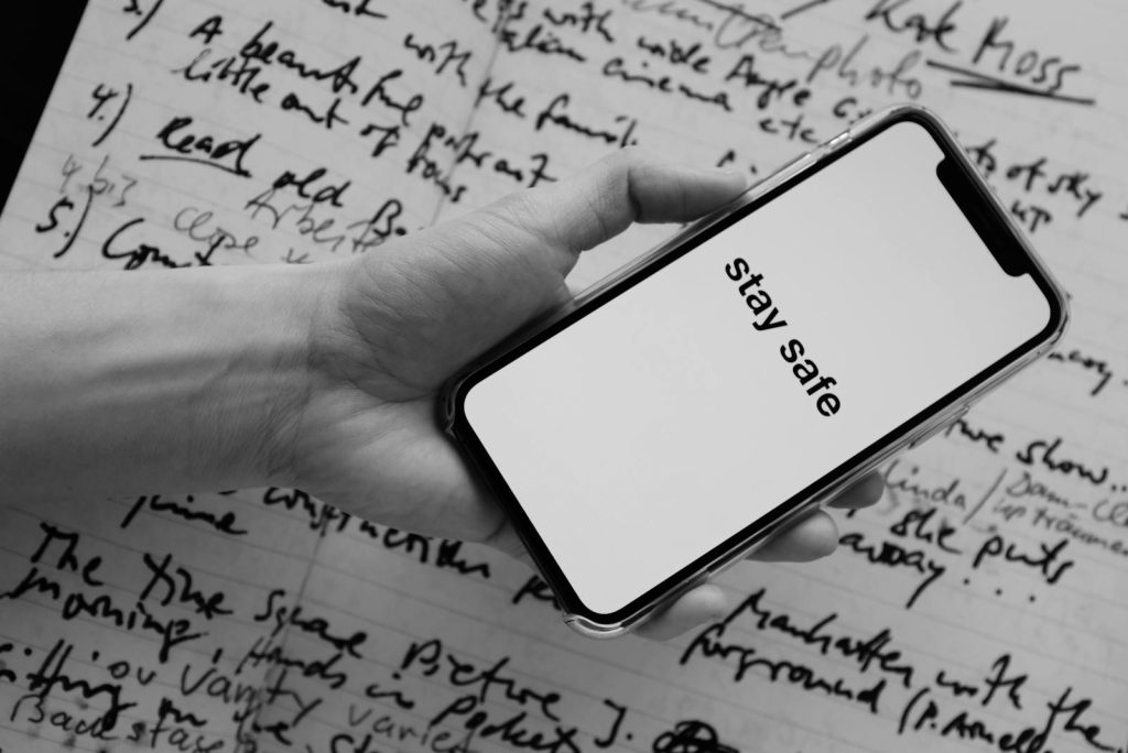 Ein Schwarz-Weiß-Bild einer Hand, die ein Smartphone mit dem Text „Bleib sicher“ hält, über handgeschriebenen Notizen.