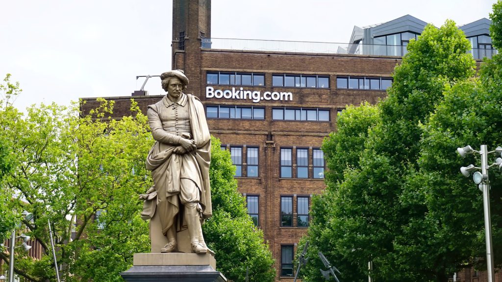 Statue eines Mannes, der tagsüber ein Buch in der Nähe eines braunen Betongebäudes hält Im Hintergrund ist das Logo von BKG Hotel Booking